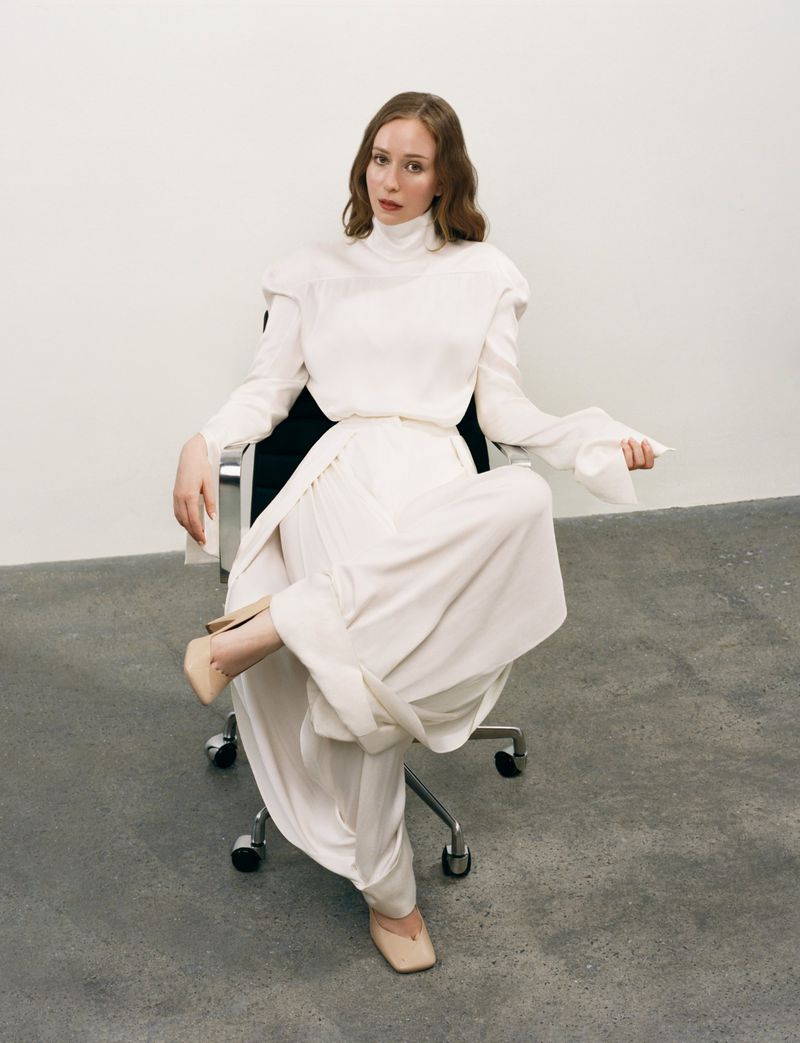 Hannah Einbinder seated in an office chair wearing an all-white flowing ensemble and beige heels against a neutral wall.