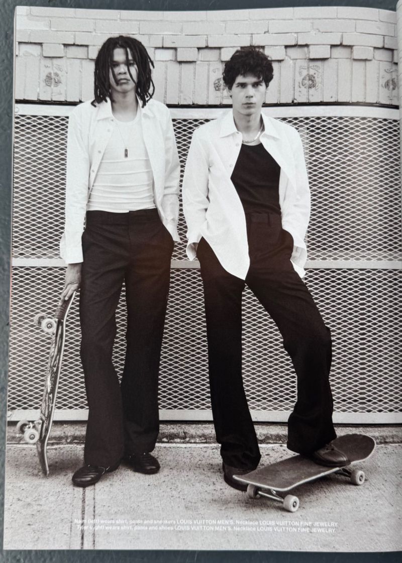 Two young adult men in unbuttoned white shirts pose outdoors with a skateboard against a concrete wall on a sunny day.