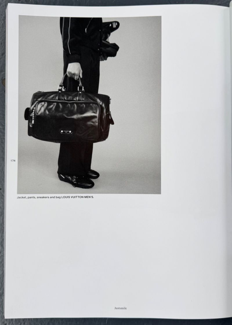 Leather duffel bag with black suit, trousers, and glossy dress shoes on a gray background, indoor studio product shot.