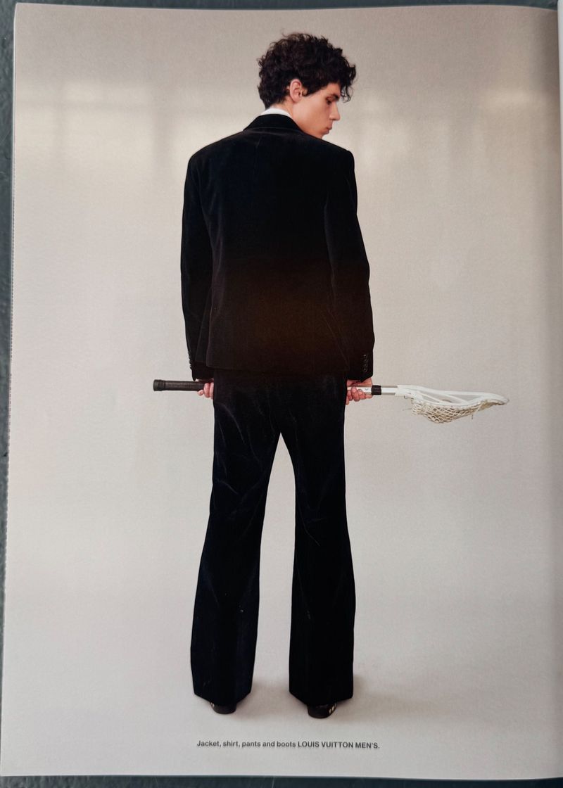 Back view of a young adult male wearing a velvet suit and holding a lacrosse stick, posing against a light gray backdrop in studio lighting.