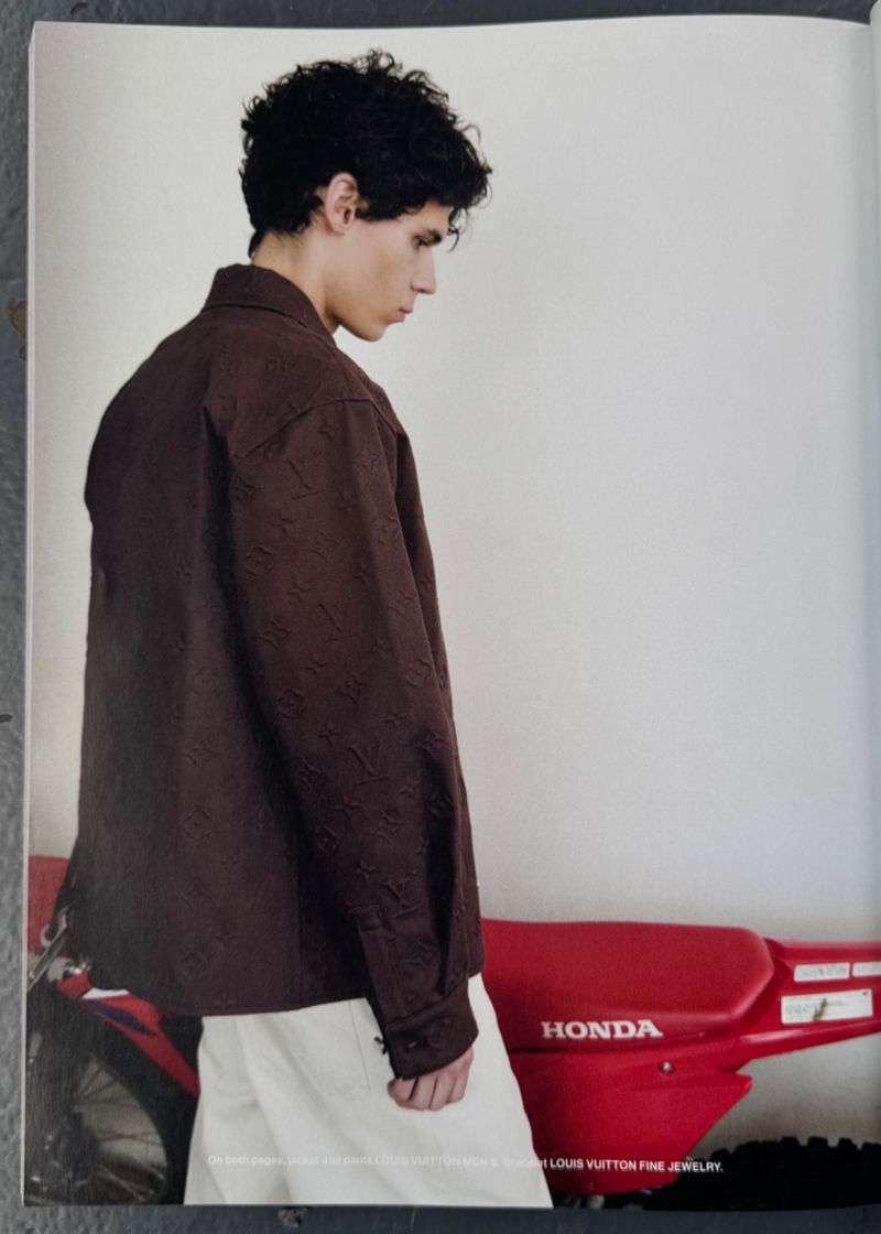 Young adult poses in a brown textured shirt with white pants beside a red Honda dirt bike against a neutral studio backdrop. 