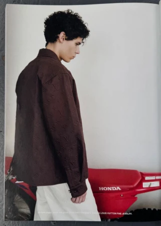 Young adult poses in a brown textured shirt with white pants beside a red Honda dirt bike against a neutral studio backdrop. 