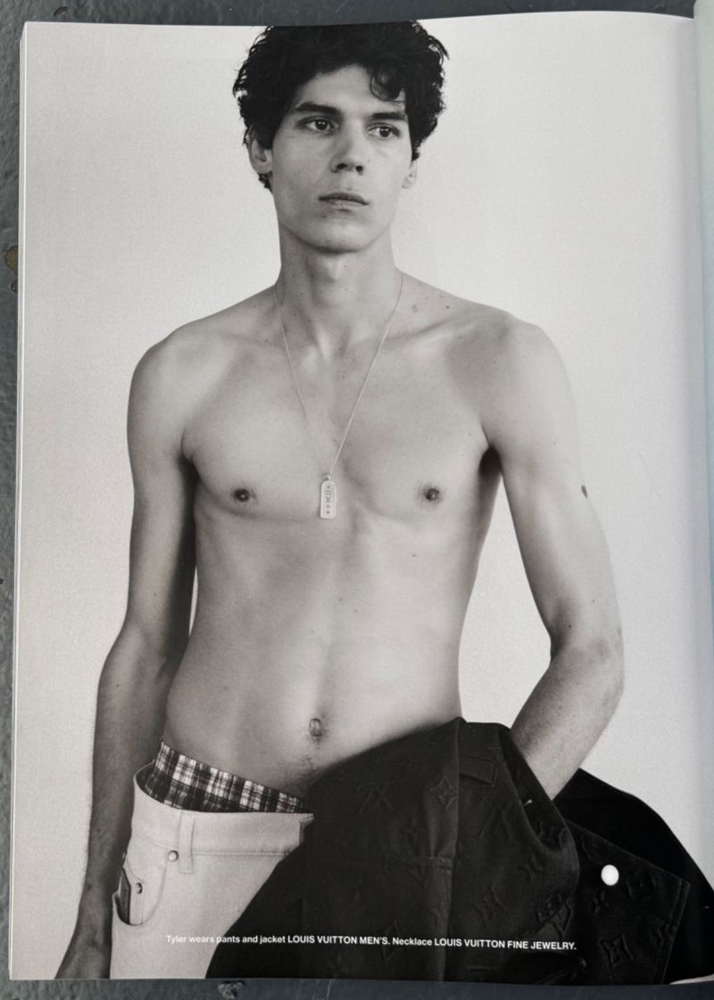 Black and white portrait of a shirtless adult male wearing a necklace, white jeans and holding a dark jacket against a light grey background.