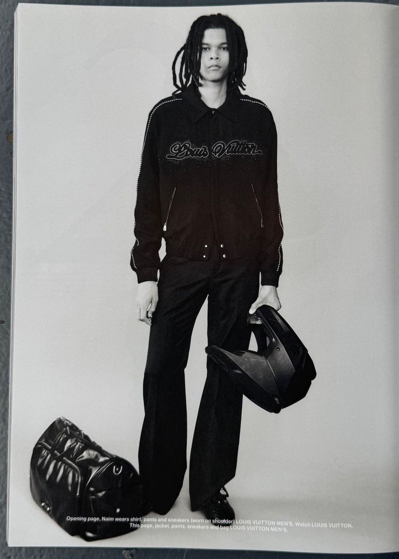 A person with dreadlocks wears a black jacket and pants while holding a helmet, with a bag on the floor in a studio shot.