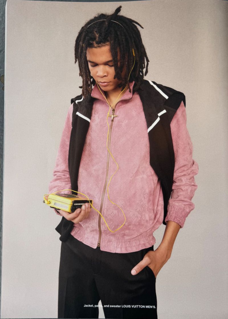 Young adult male with dreadlocks wearing a pink jacket and black vest, listening to a yellow walkman against a grey background.
