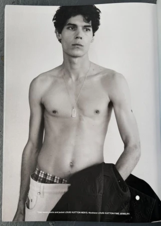 Shirtless young man wearing white trousers and holding a dark jacket against a plain background