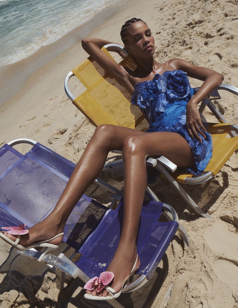 Model in a textured blue strapless mini dress reclining on beach chairs on a sunny sandy beach.
