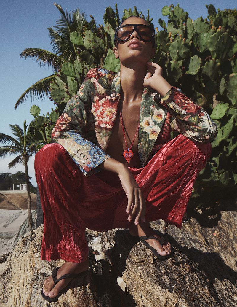 Fashion model crouching on a rock wearing shield sunglasses, a floral print blazer, and shiny red trousers in a tropical cactus setting.