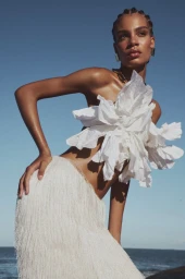 Model wearing a white sculptural flower top and fringed skirt posing outdoors against a clear blue sky and ocean.