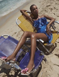 Model in a textured blue strapless mini dress reclining on beach chairs on a sunny sandy beach.