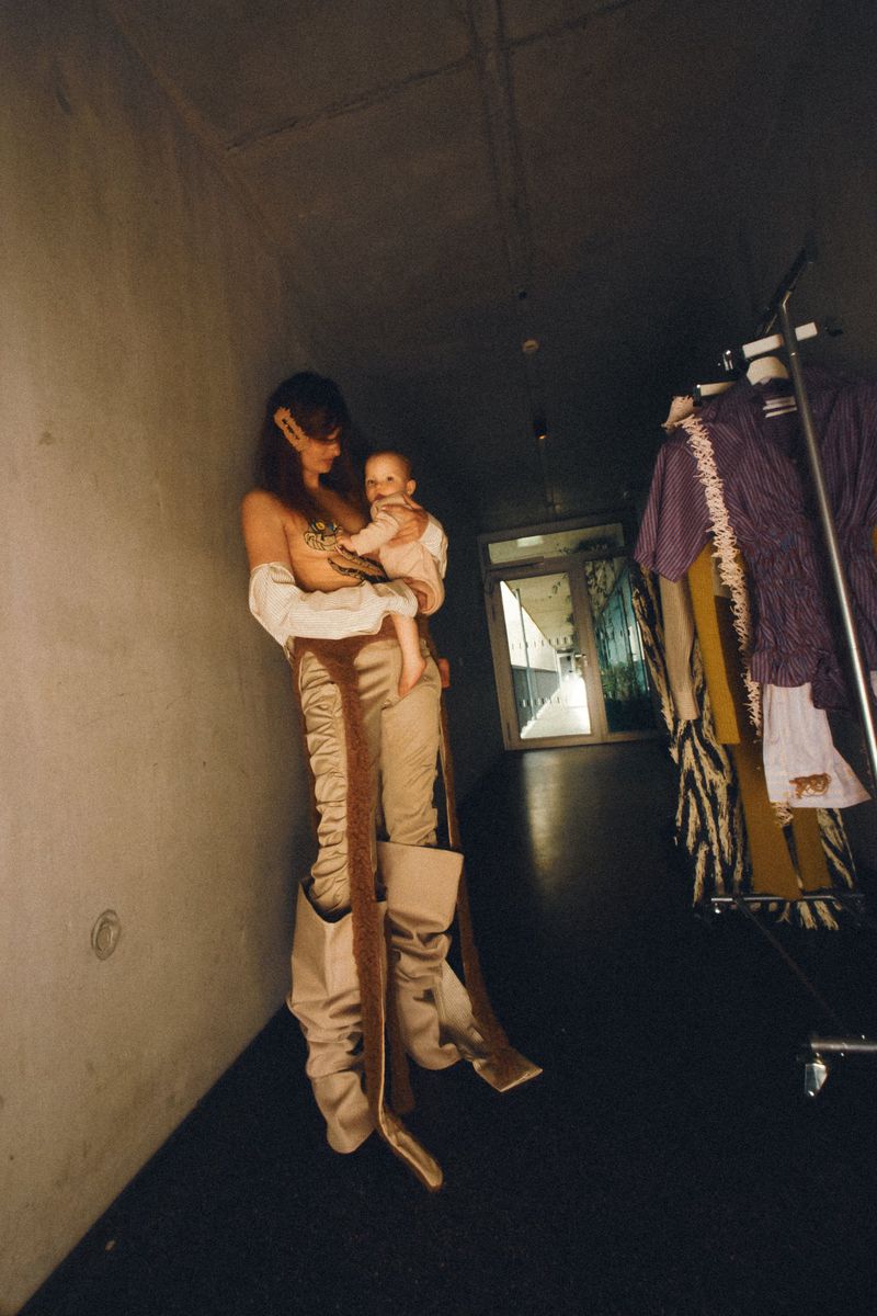 A woman holding an infant stands in a dim hallway wearing avant-garde ruched beige trousers and exaggerated slouchy boots.