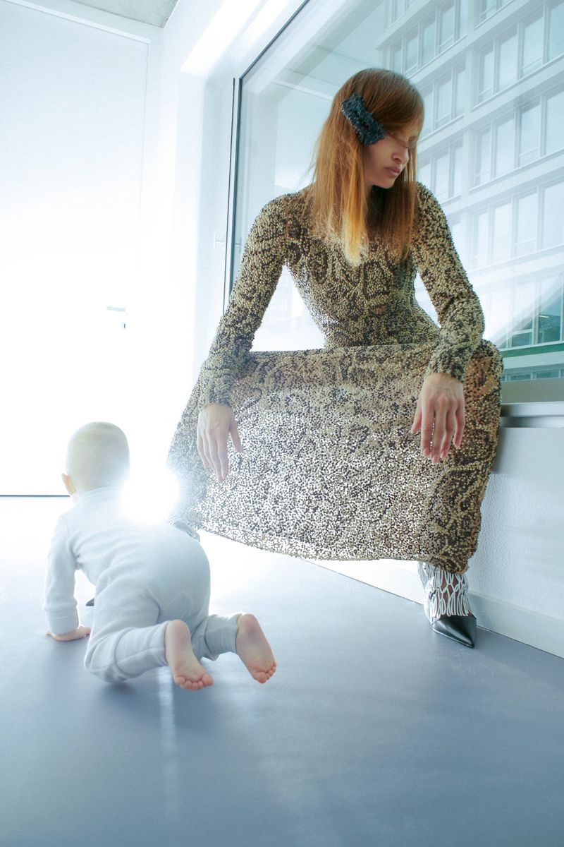 Woman in long snakeskin-pattern dress crouching by a window while a baby crawls on the light grey floor below