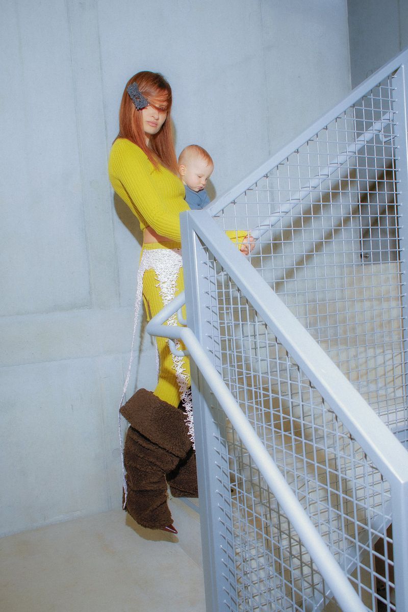 Woman with auburn hair in a yellow knit set and oversized brown boots holds a baby on an industrial staircase behind a metal mesh railing.