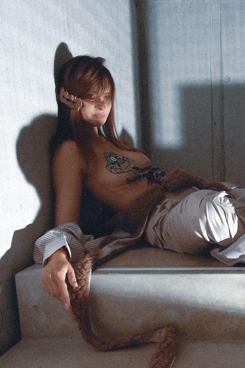 Woman with chest artwork and brown faux fur trim reclining on concrete steps beside a concrete wall in directional lighting.