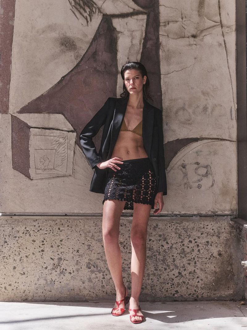 Adult woman in a blazer, top and black lace skirt, posing against a patterned wall in an outdoor environment, fashion editorial.
