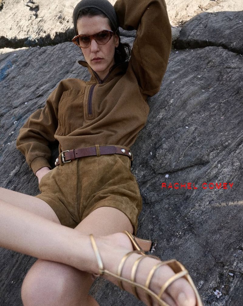 A woman poses outdoors wearing a brown pullover, matching shorts, sunglasses, beanie, studded belt, and gold strappy sandals.