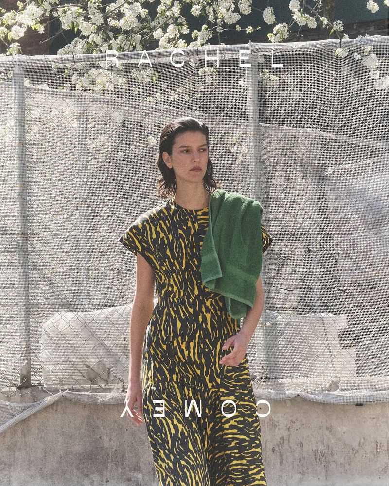 A brunette woman with a towel over her shoulder stands outside, wearing a patterned black and yellow dress in this fashion shot.