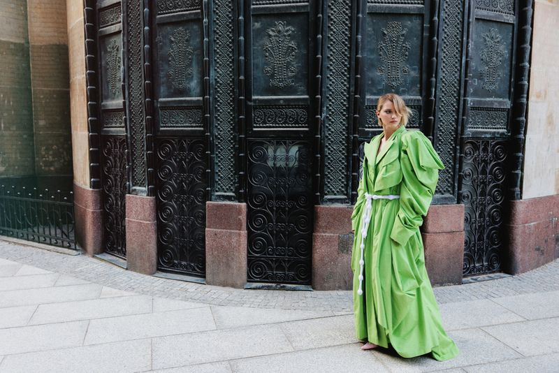 Model Daga Jez in an oversized bright green belted coat standing outside ornate dark iron architectural doors on stone pavement.