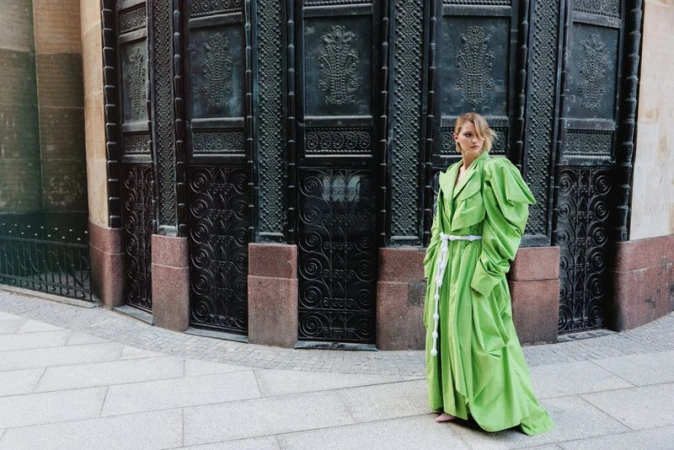 Model Daga Jez in an oversized bright green belted coat standing outside ornate dark iron architectural doors on stone pavement.