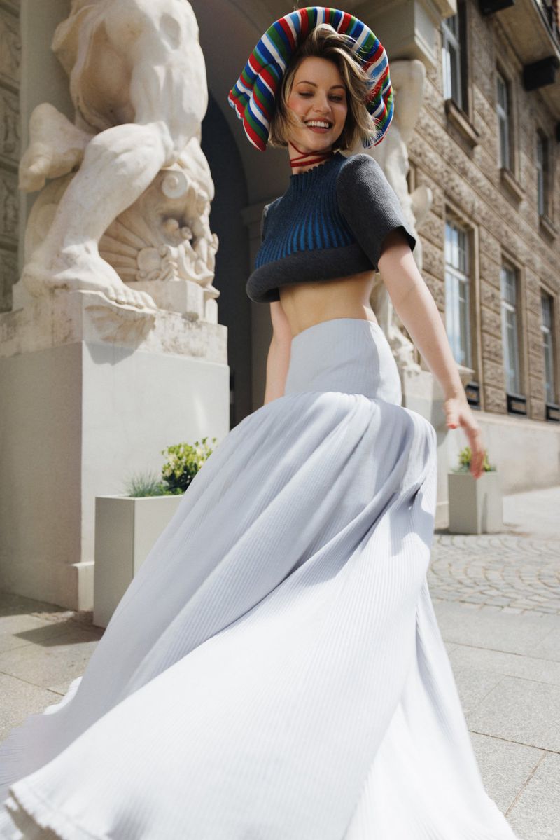 Model Daga Jez smiling outdoors wearing a voluminous white maxi skirt, cropped top, and colorful headpiece by classical statues.