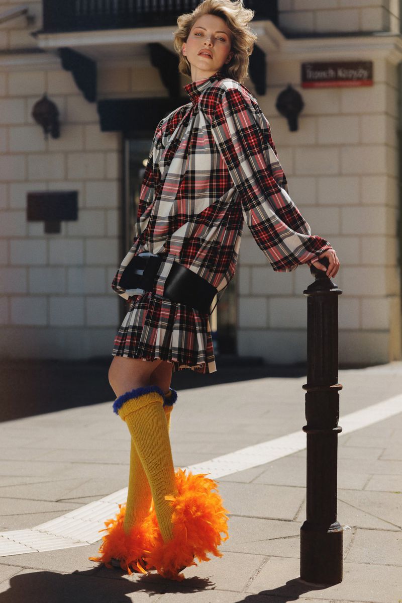 Model Daga Jez in a red and white plaid dress with yellow socks and orange feathered shoes leaning on a street bollard.