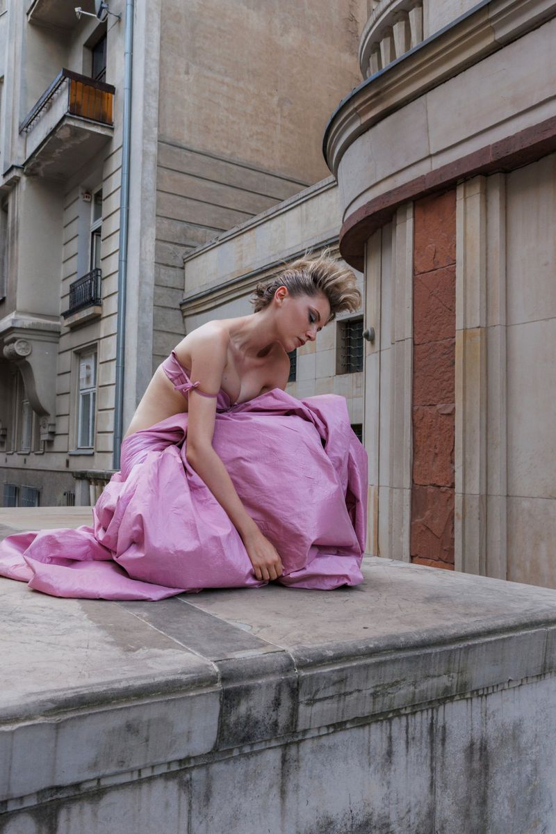 Model Daga Jez crouching on an architectural stone ledge wearing a voluminous pink gown against historic building facades.