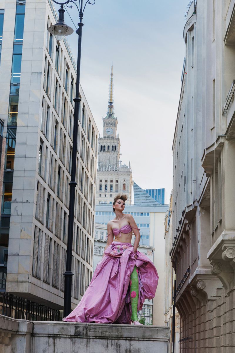 Model Daga Jez in a voluminous pink gown and green boots posing on an architectural ledge between city buildings.