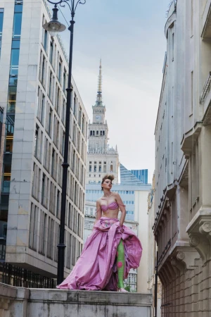 Model Daga Jez in a voluminous pink gown and green boots posing on an architectural ledge between city buildings.