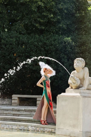 Model Daga Jez wearing a patterned gown and white feathered hat posing next to a stone cherub water fountain outdoors.