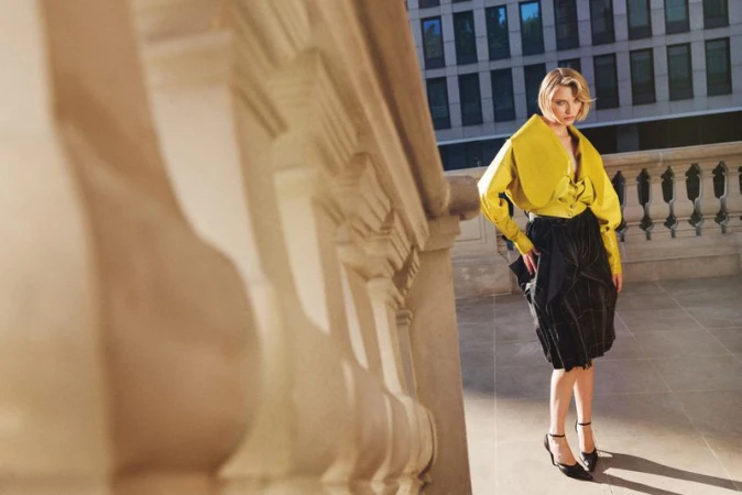 Model Daga Jez wearing a bright yellow jacket and pleated black skirt posing on a terrace framed by a stone balustrade.