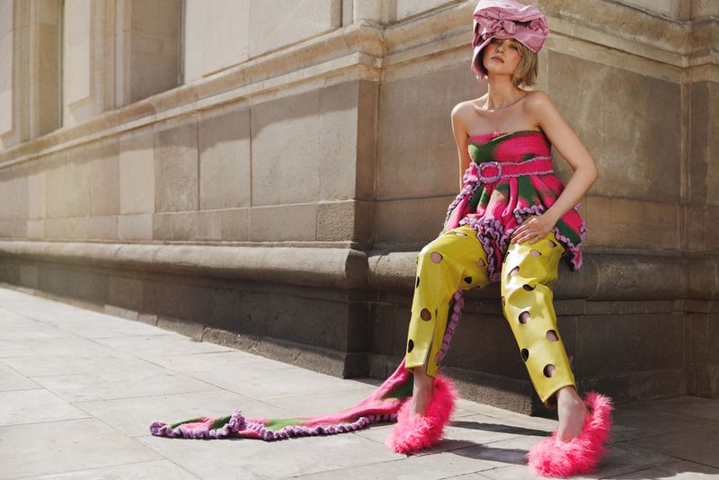 Model Daga Jez seated on a stone building ledge wearing a pink headpiece, cutout yellow trousers, and fuzzy pink slippers.