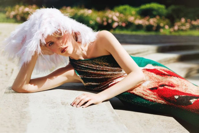 Model Daga Jez reclining on stone steps in a vibrant knit dress and oversized white feather hat for Elle Kazakhstan.