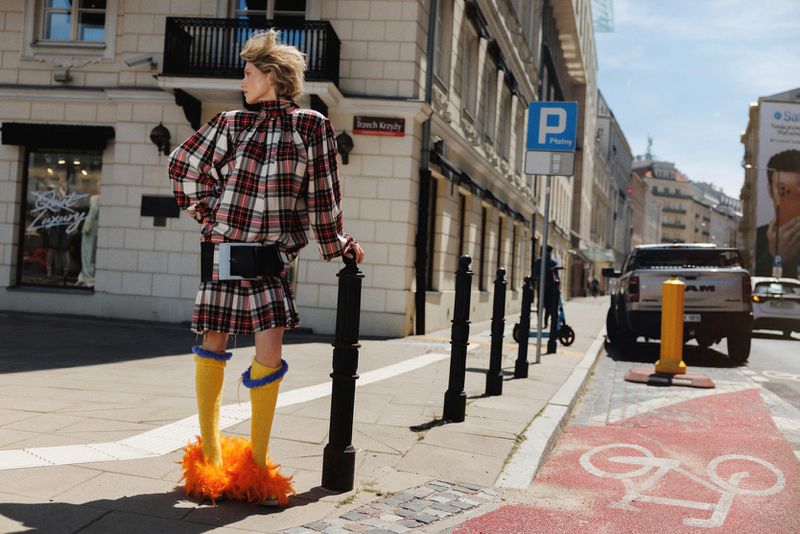 Model in a plaid skirt suit and bright yellow knee socks with orange feather cuffs standing on a European city street