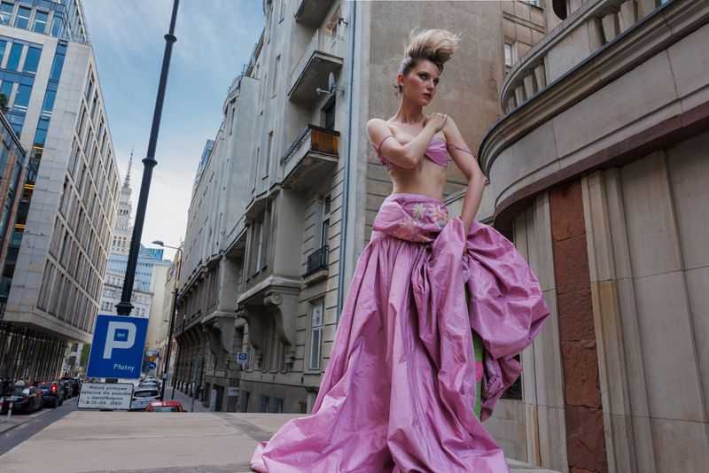 Model wearing a voluminous pink gown standing outdoors on a city street between tall historic buildings.