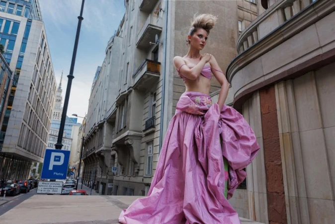 Model wearing a voluminous pink gown standing outdoors on a city street between tall historic buildings.