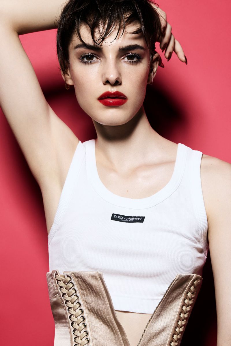 Model Carla Rey with short dark hair and bold red lips wearing a white tank top and lace-up corset against a bright pink background.