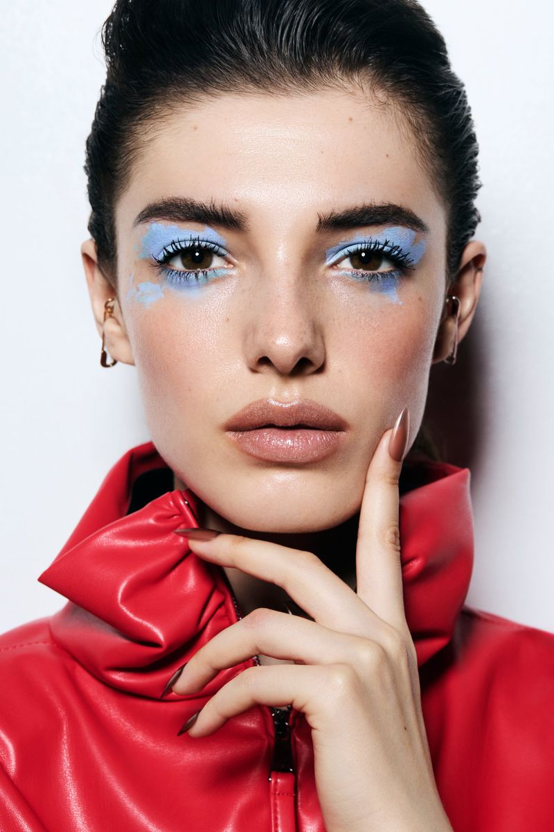 Model Carla Rey with vivid blue eye makeup and a high-collar red leather jacket touching her chin against a white studio backdrop.