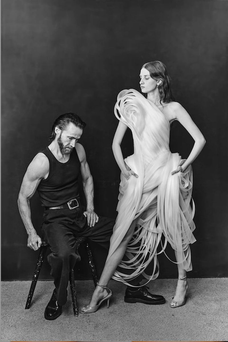 Woman in a sculptural white ribbon dress standing beside a seated bearded man in a black tank top against a dark studio backdrop.