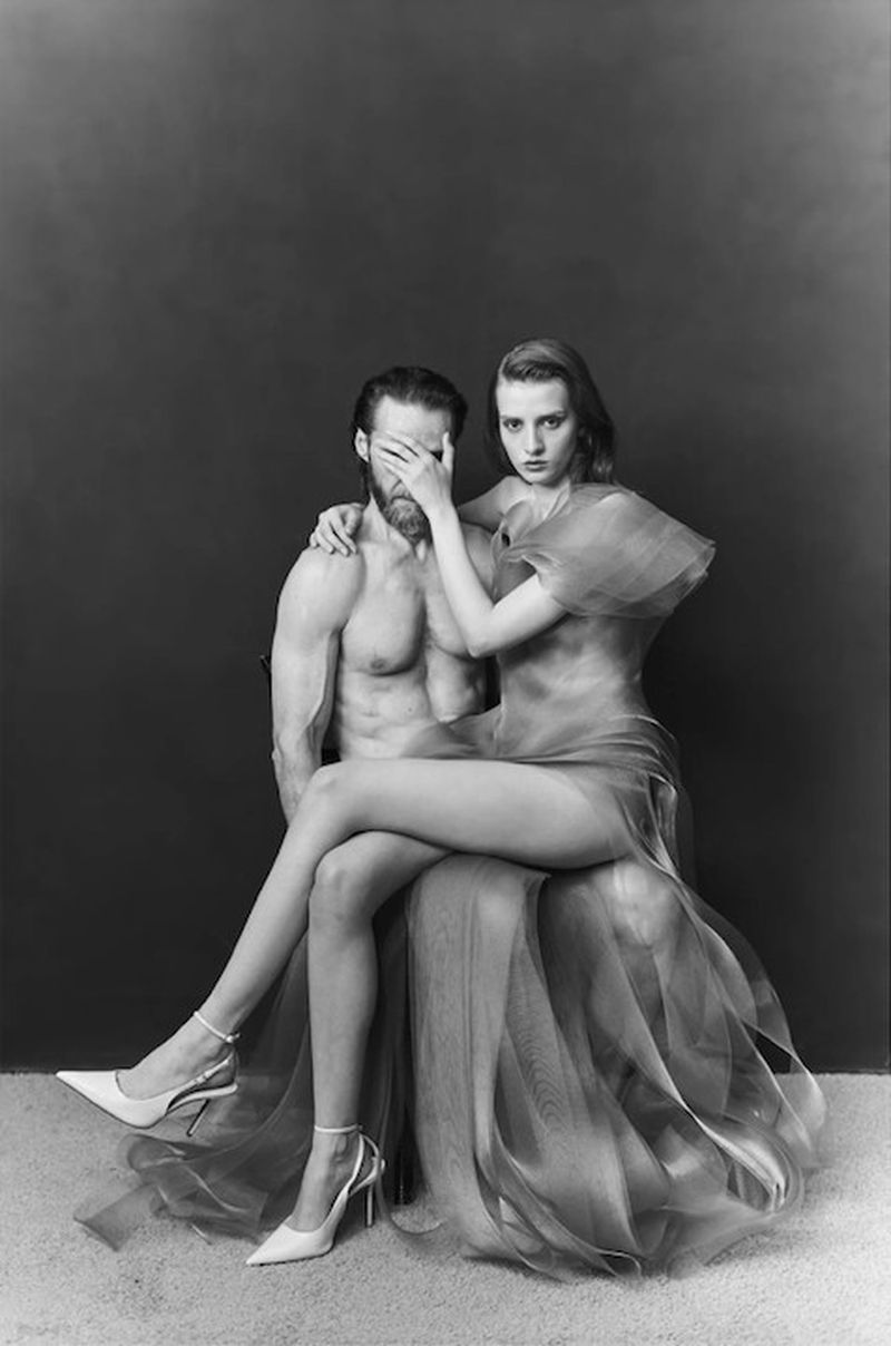 Black and white editorial portrait of a woman in a sheer gown covering a shirtless bearded man's eyes while seated on his lap.