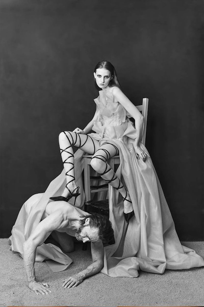 Monochrome editorial portrait of a woman seated on a wooden chair resting her lace-up stiletto heel on the back of a crawling man.