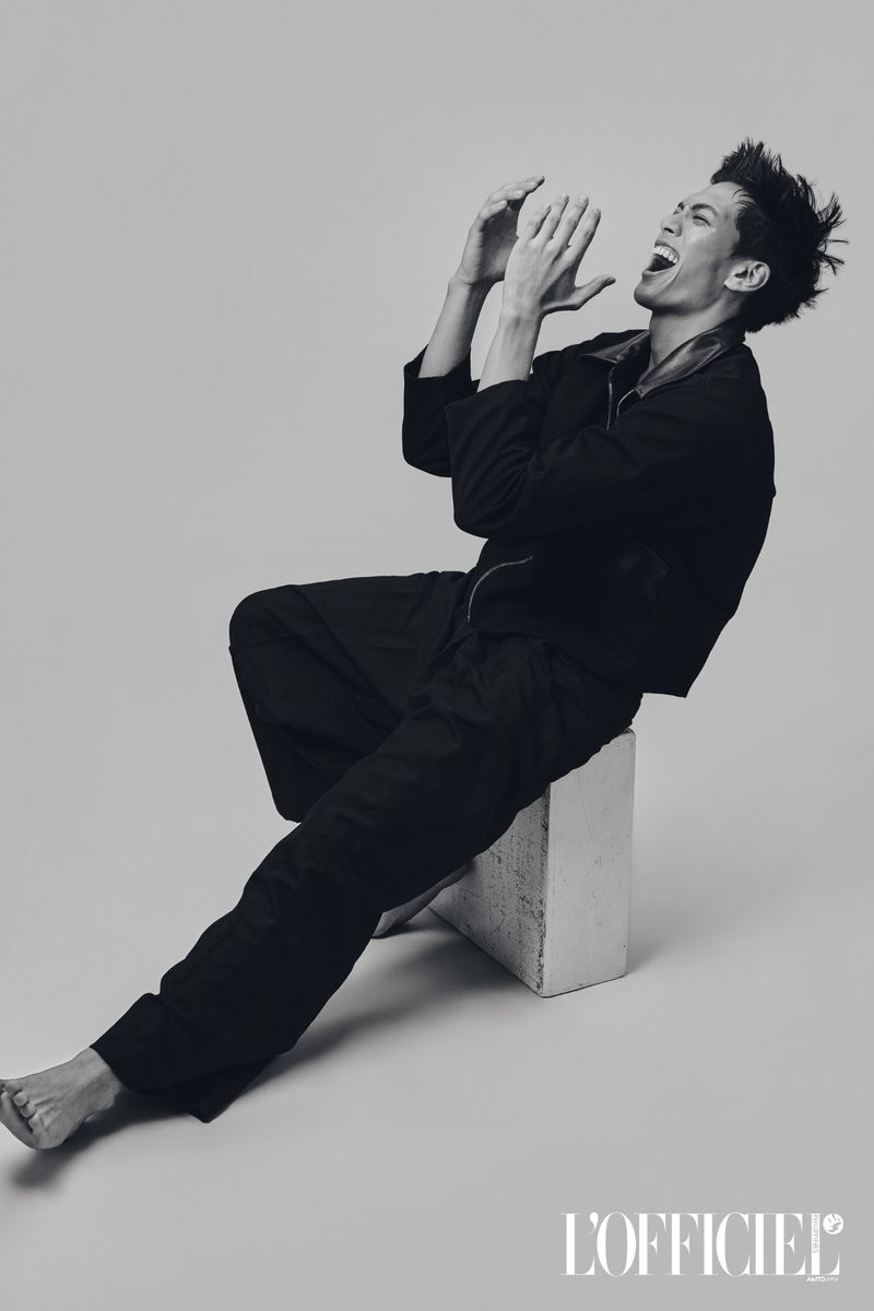 Barefoot man in an all-black outfit sitting on a white block while laughing in a monochrome studio setting.