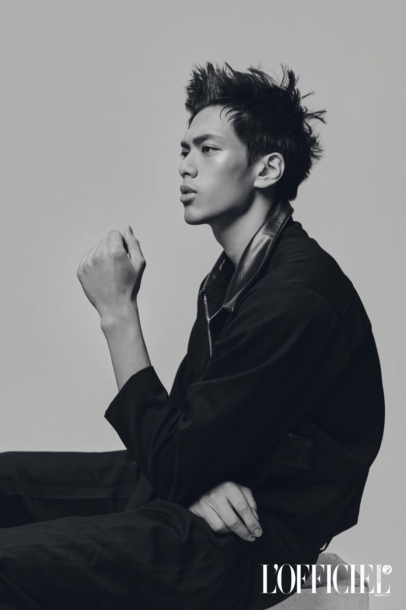 Side profile portrait of a young man with styled spiky hair in a black collared jacket against a neutral studio backdrop.