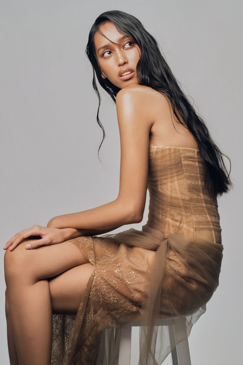 A seated female model wearing a sheer brown strapless dress against a neutral grey studio background
