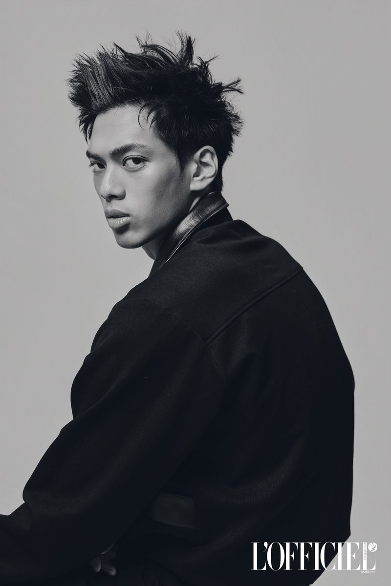 Male model with spiked dark hair wearing a black jacket looking over his shoulder against a plain grey studio backdrop.