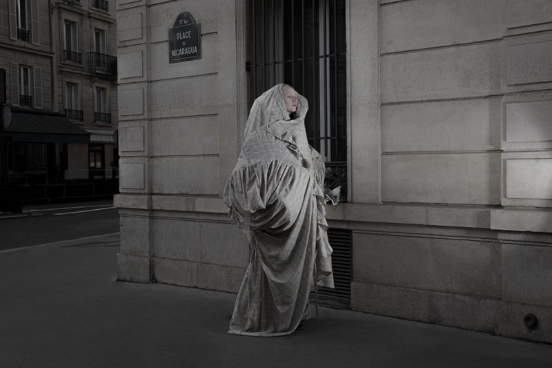 Model draped in heavy patterned fabric standing on a street corner against a classical stone building facade in a monochrome photograph.