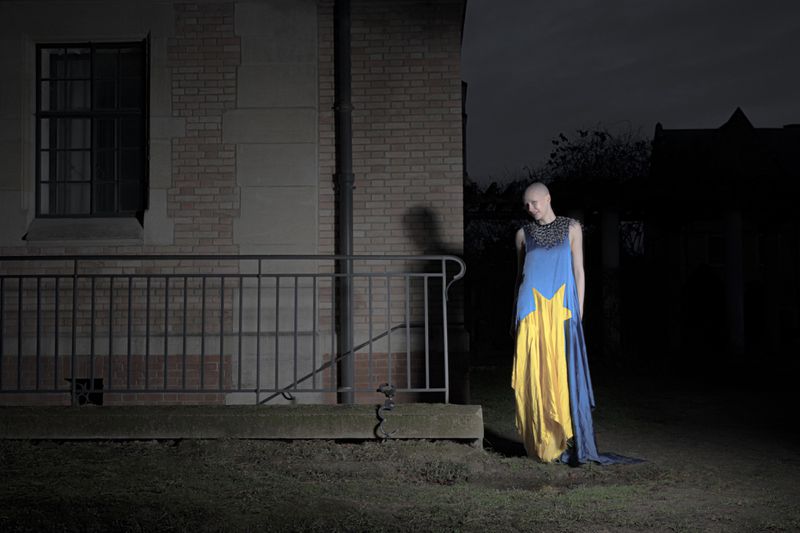 Model with a shaved head in a blue and yellow star-patterned gown standing beside a brick building at night.