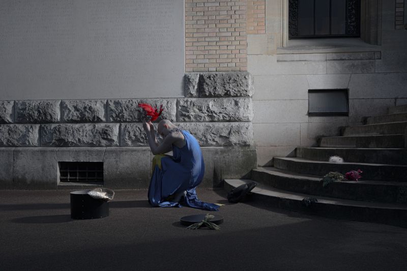 Model Victoria Cassie kneeling outdoors in a blue draped dress holding red feathers near stone architectural steps.