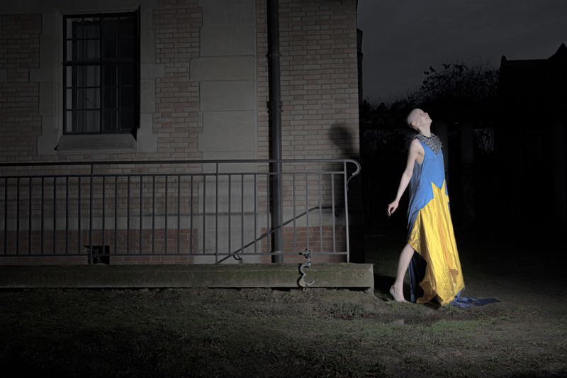 Model with a shaved head in a blue and yellow dress looking upward at night outside a brick building with a metal railing.