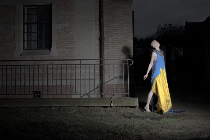 Model with a shaved head in a blue and yellow dress looking upward at night outside a brick building with a metal railing.