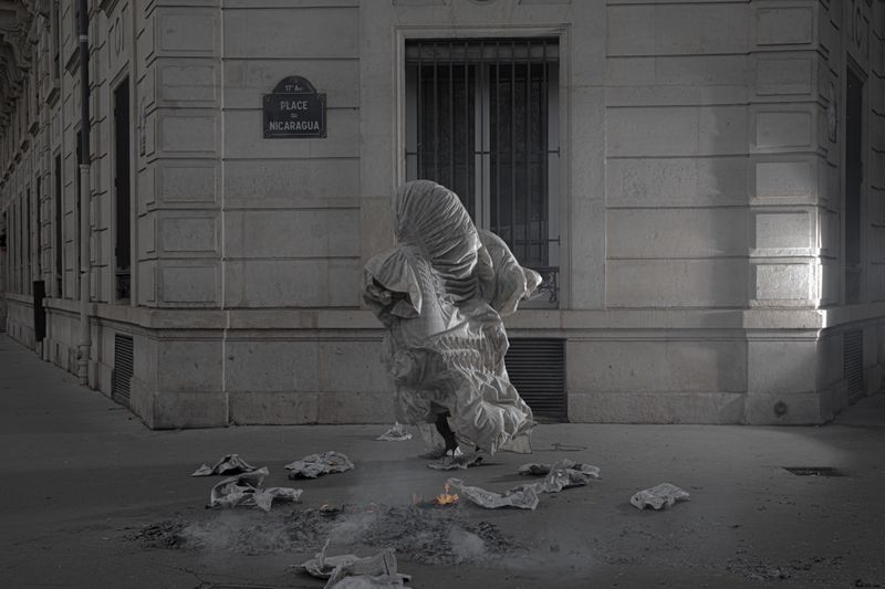 Model completely covered in a voluminous draped white gown standing on a street corner near scattered paper embers.